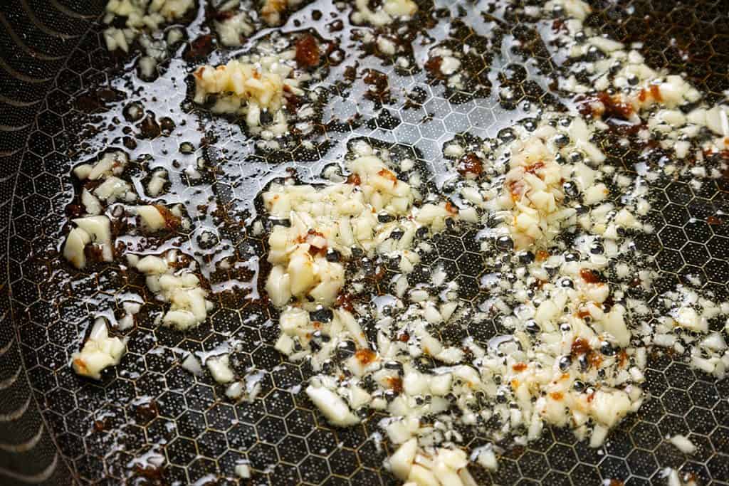 Chopped garlic sizzling in oil on a black hexagonal-patterned pan, starting to brown&mdash;the perfect aromatic base for Baked Garlic Chicken Thighs.