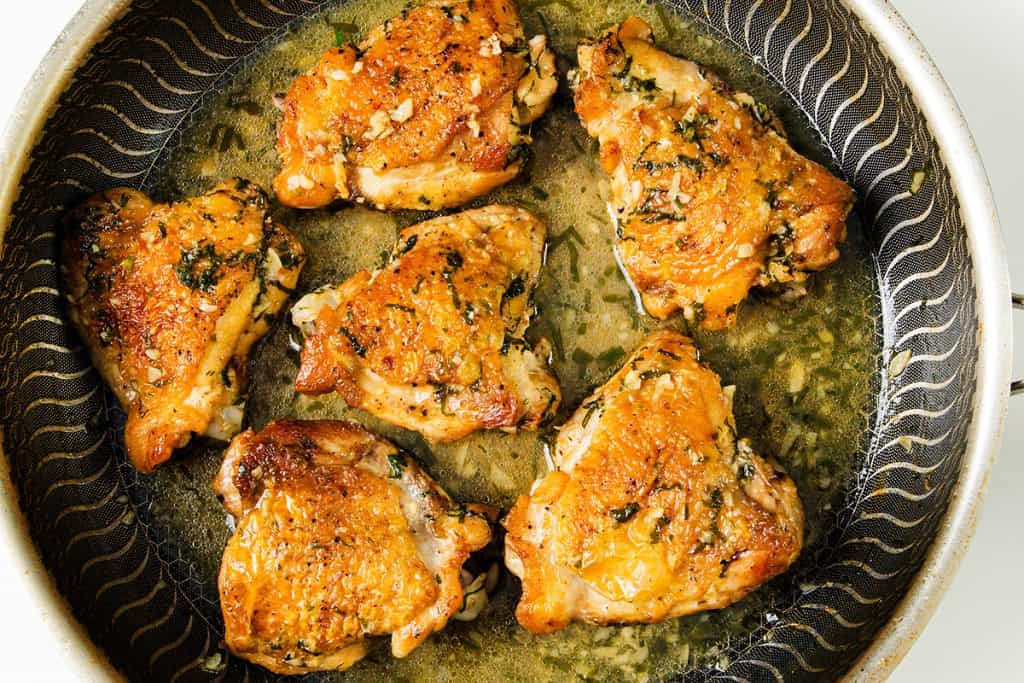 Six golden brown Baked Garlic Chicken Thighs with crispy skin and sprinkled herbs are cooking in a frying pan, with a bit of oil and minced garlic visible around them.