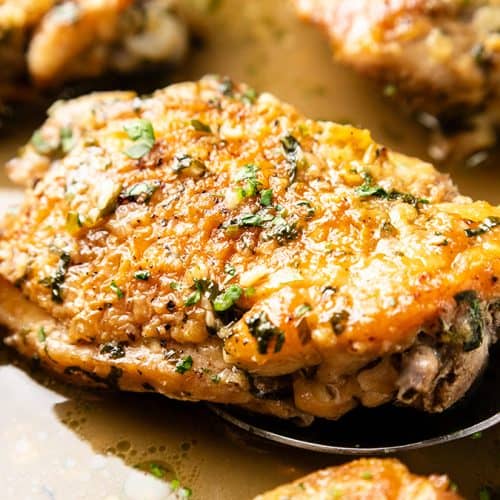 Close-up of a spoon lifting a piece of golden-brown, herb-seasoned Baked Garlic Chicken Thigh in a pan with sauce. The chicken looks crispy and juicy, garnished with herbs, and surrounded by more pieces in the background.