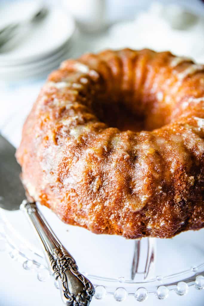 An Orange Bundt cake with a sugar glaze on a glass cake stand with a cake slicer next to it