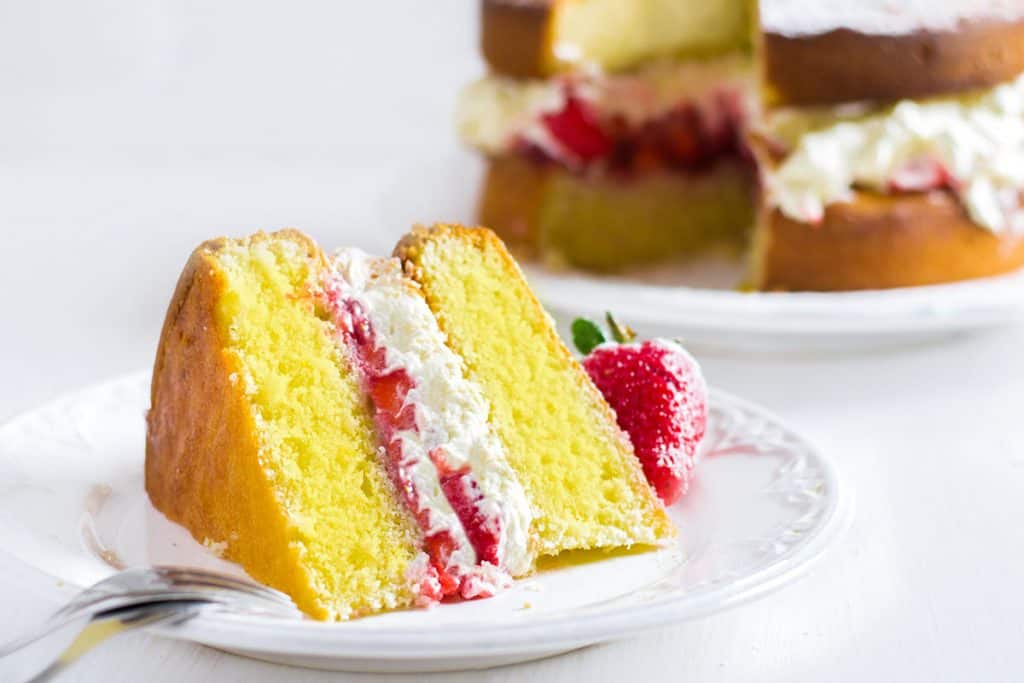A slice of Lemon Sponge Cake filled with whipped cream and strawberries on a plate with the cake behind it