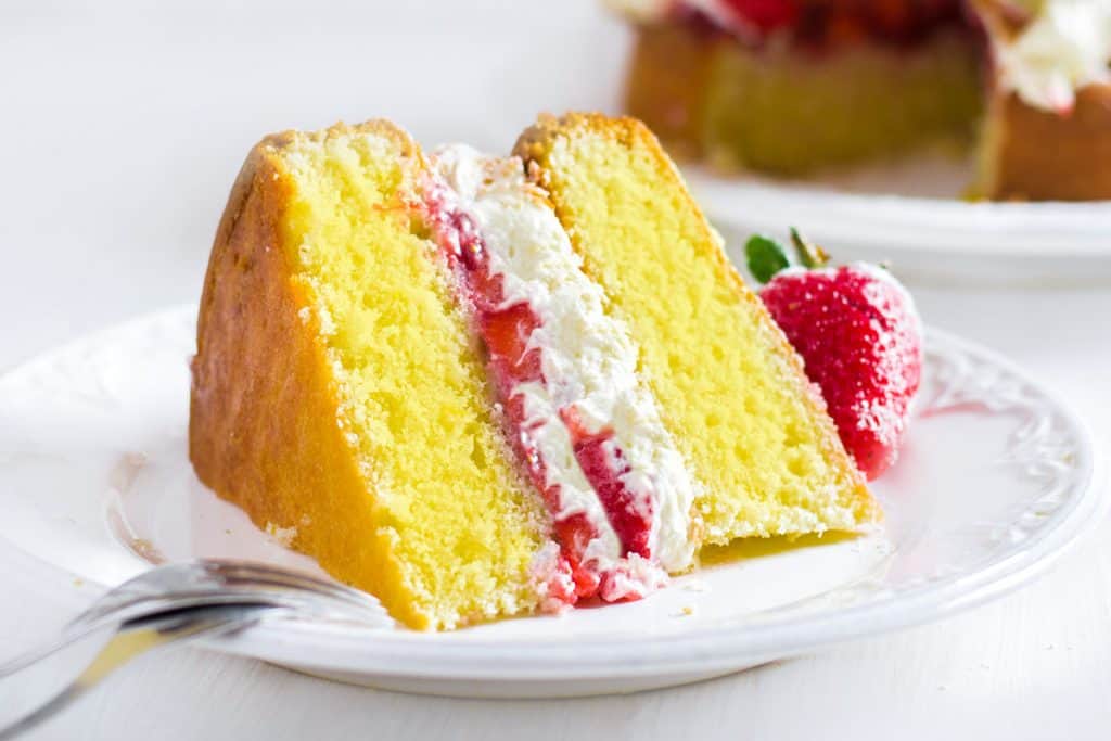 A slice of Lemon Sponge Cake filled with whipped cream and strawberries on a plate with the cake behind it