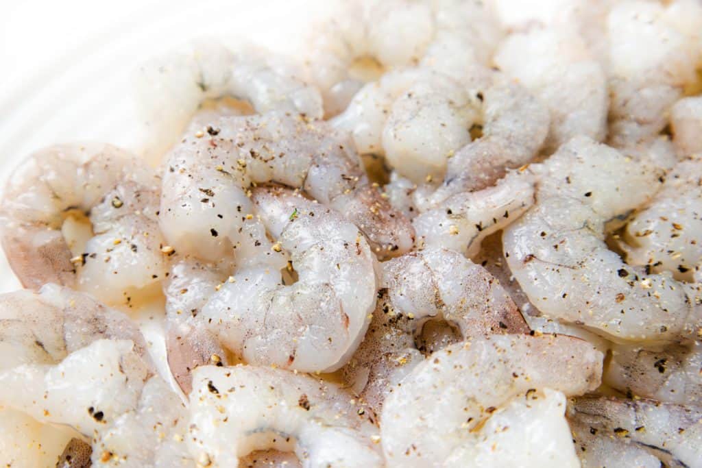 Raw, deveined shrimp seasoned with salt and cracked black pepper, arranged closely together on a white surface—perfectly prepped for a delicious Coconut Shrimp Curry.
