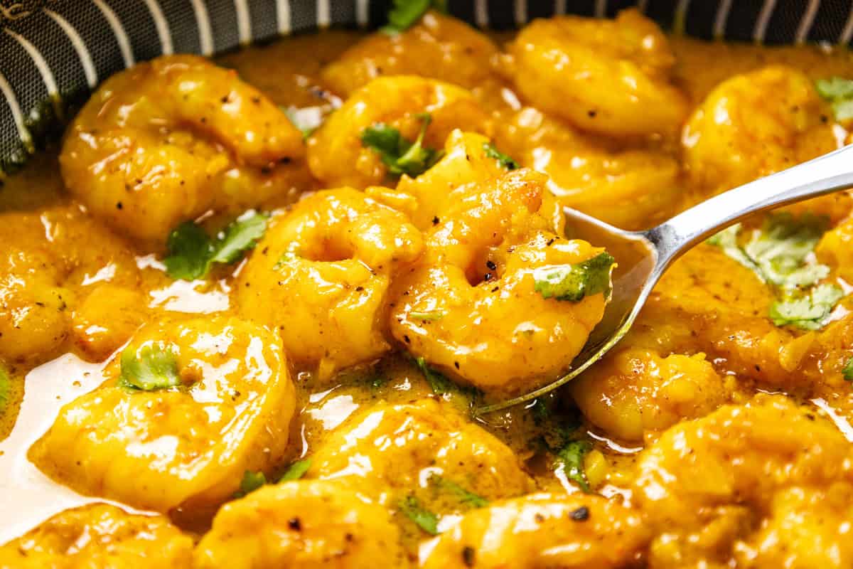 Close-up of Coconut Shrimp Curry cooked in a rich, yellow curry sauce with herbs, served in a bowl. A spoon is lifting some shrimp, highlighting the creamy texture and garnished with chopped green herbs.