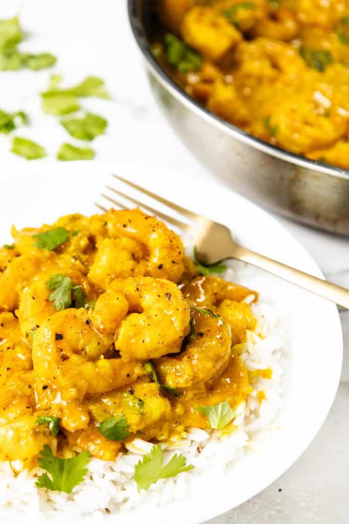 A white plate of Coconut Shrimp Curry with rice garnished with cilantro, with a gold fork beside it. In the background, a pan filled with more shrimp curry is partially visible.