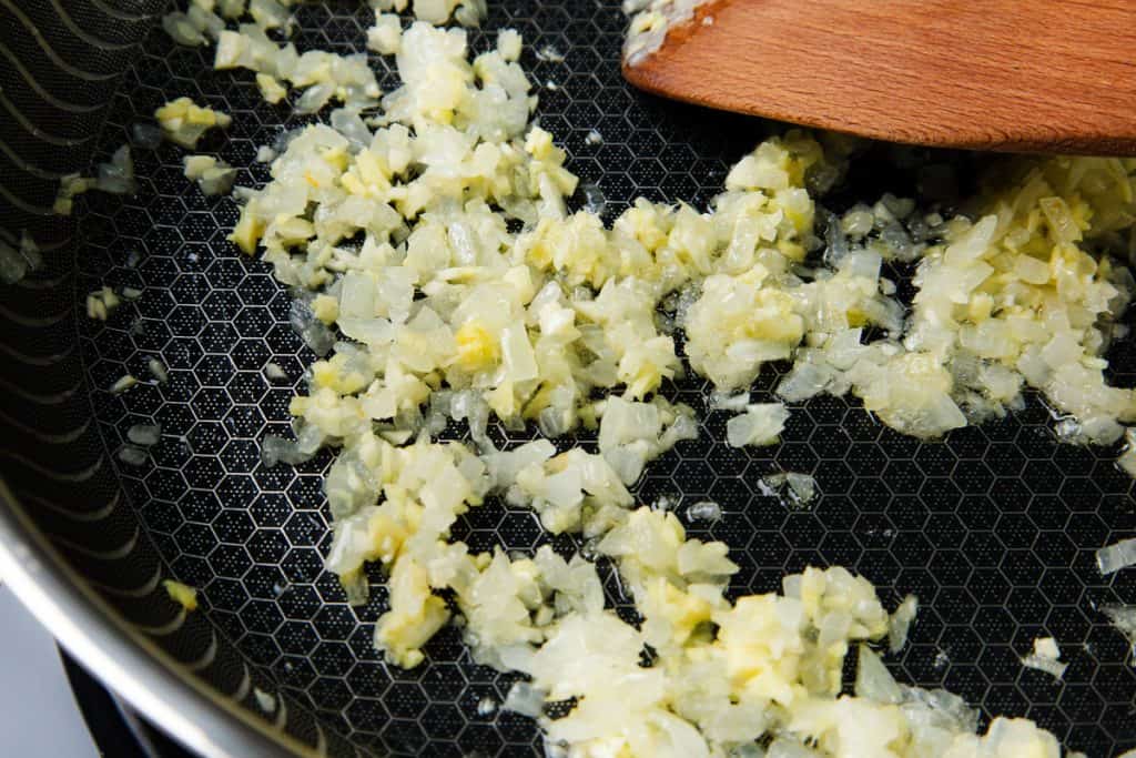 Chopped onions and garlic sautéing in a frying pan, being stirred with a wooden spatula—the perfect aromatic base for a flavorful Coconut Shrimp Curry.