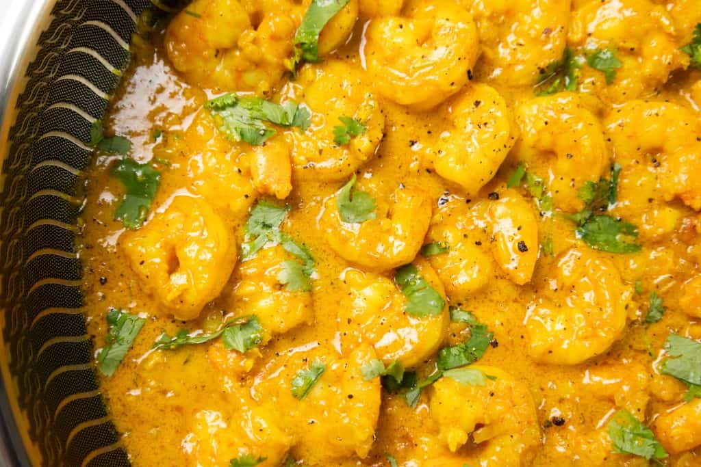 Close-up of Coconut Shrimp Curry cooked in a yellow curry sauce, garnished with chopped cilantro and ground black pepper. The shrimp are coated in a thick, creamy sauce with visible herbs and spices.