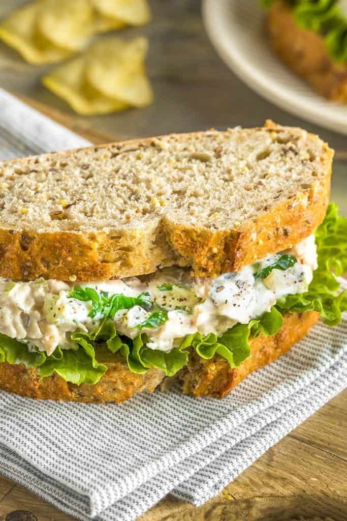 A tuna salad sandwich on a cutting board with potato chips in the background