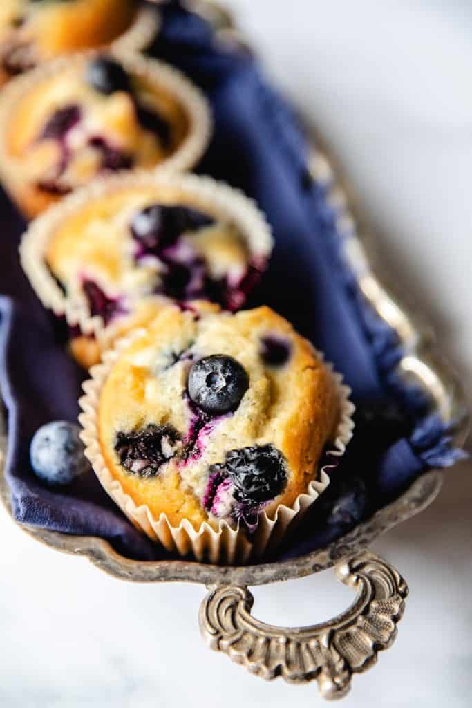 Five Blueberry muffins lined up on a silver tray with an ornate handle