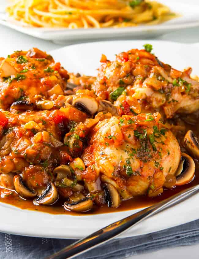 a serving dish full of Chicken Cacciatore with a plate of spaghetti in the background.