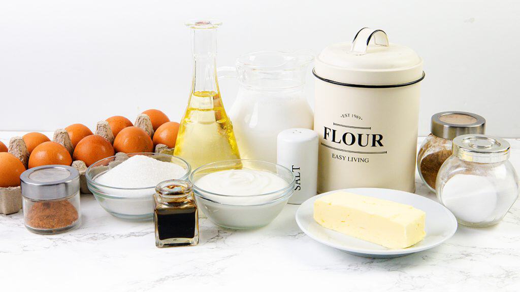 A variety of baking ingredients on a white countertop, including eggs, flour, sugar, salt, butter, oil, milk, vanilla extract, cocoa powder, and a bowl of yogurt or sour cream—perfect for making delicious Sour Cream Pancakes.