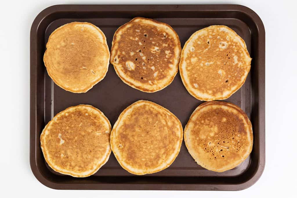 Six golden-brown sour cream pancakes arranged in two rows of three on a dark baking tray, viewed from above on a white background.
