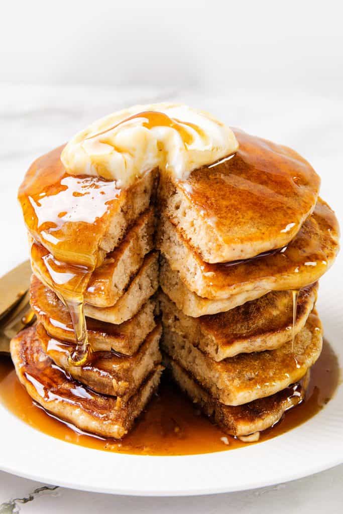 A stack of sour cream pancakes with a pat of melting butter and maple syrup drizzling down, served on a white plate. A wedge has been cut out, revealing the fluffy texture inside.