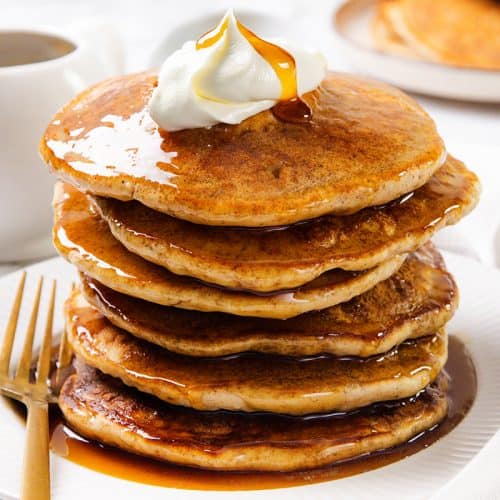 A stack of fluffy sour cream pancakes topped with whipped cream and syrup on a white plate, with a gold fork on the side and a syrup pitcher and more pancakes in the background.