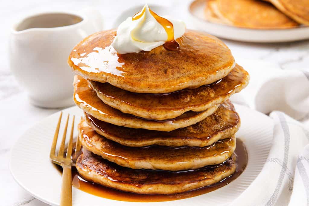 A stack of fluffy sour cream pancakes topped with whipped cream and syrup sits on a white plate with a gold fork. A small pitcher of syrup and another plate of pancakes can be seen in the background.