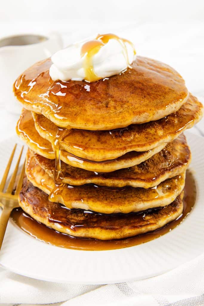 A stack of golden brown sour cream pancakes topped with a dollop of whipped cream and drizzled with syrup, served on a white plate with a fork and a cup of coffee in the background.