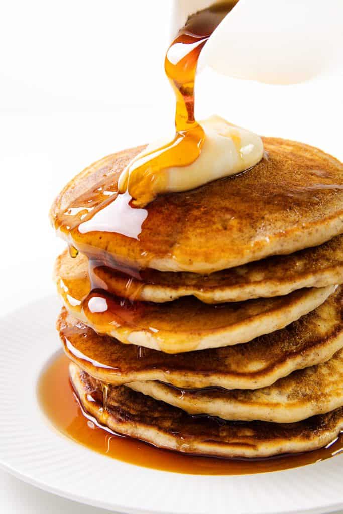 A stack of five sour cream pancakes topped with a pat of butter, with syrup being poured over them, on a white plate against a white background.