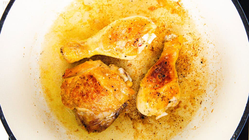 Three pieces of golden-brown roasted chicken, reminiscent of a rustic Coq Au Vin, include two drumsticks and one breast piece in a white pan with seasoned oil and crispy bits at the bottom.