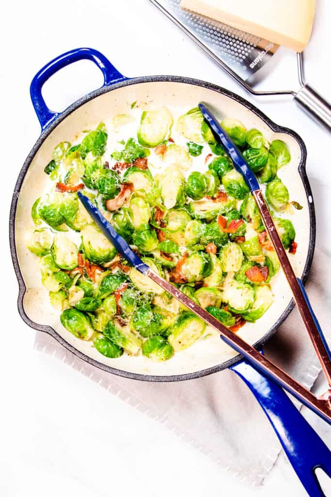a pan with brussels sprouts and bacon in a cream sauce and a pair of tongs in the pan
