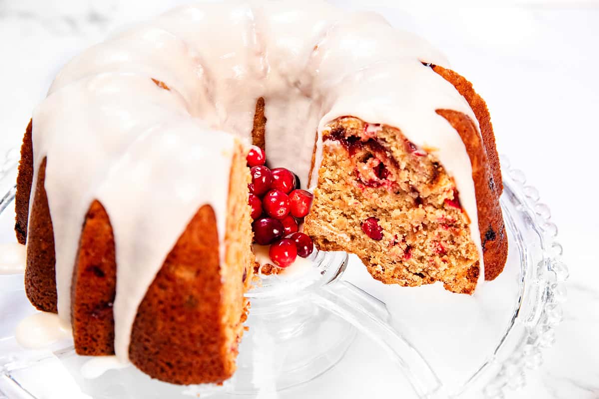 A Cranberry Christmas Bundt cake with a slice removed is displayed on a glass stand. The cake, topped with white icing, reveals a texture speckled with red berries inside. A cluster of fresh red berries elegantly decorates the center.