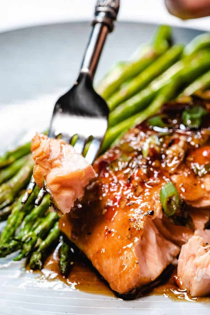 A fork with salmon on it. A plate of salmon and asparagus in the background.