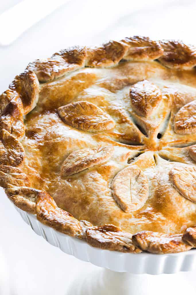 A golden-brown pie with a braided crust and decorative leaf-shaped pastry pieces on top, displayed on a white pedestal stand, showcases the beauty of learning how to make pie crust from scratch.