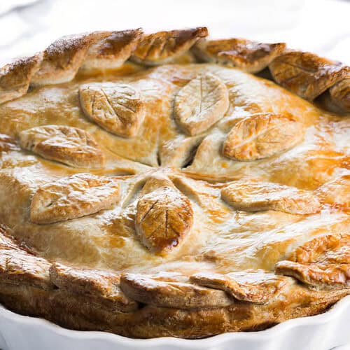A golden-brown pie with a decorative crust—showcasing leaf-shaped pastry designs made using classic how to make pie crust techniques—sits in a white ceramic dish on a light marble surface.