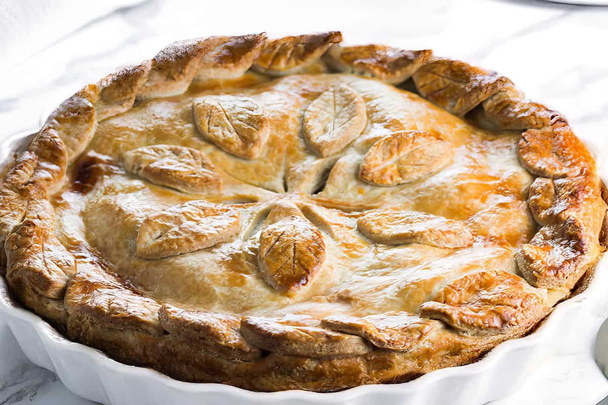 A golden-brown pie with a decorative crust—showcasing leaf-shaped pastry designs made using classic how to make pie crust techniques—sits in a white ceramic dish on a light marble surface.