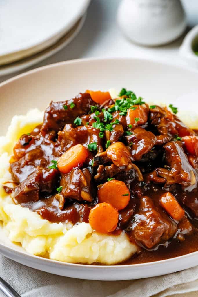 A dish of beef stew with chunks of braised boneless short ribs, carrots, and a rich brown gravy served over a bed of creamy mashed potatoes.