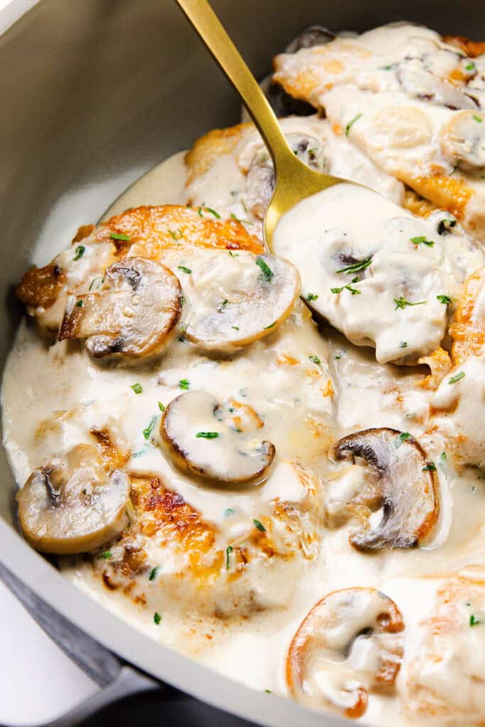 Close-up of golden-brown chicken breasts in a creamy mushroom sauce with sliced mushrooms, garnished with chopped herbs, in a pan with a gold spoon scooping up some of the flavorful sauce.