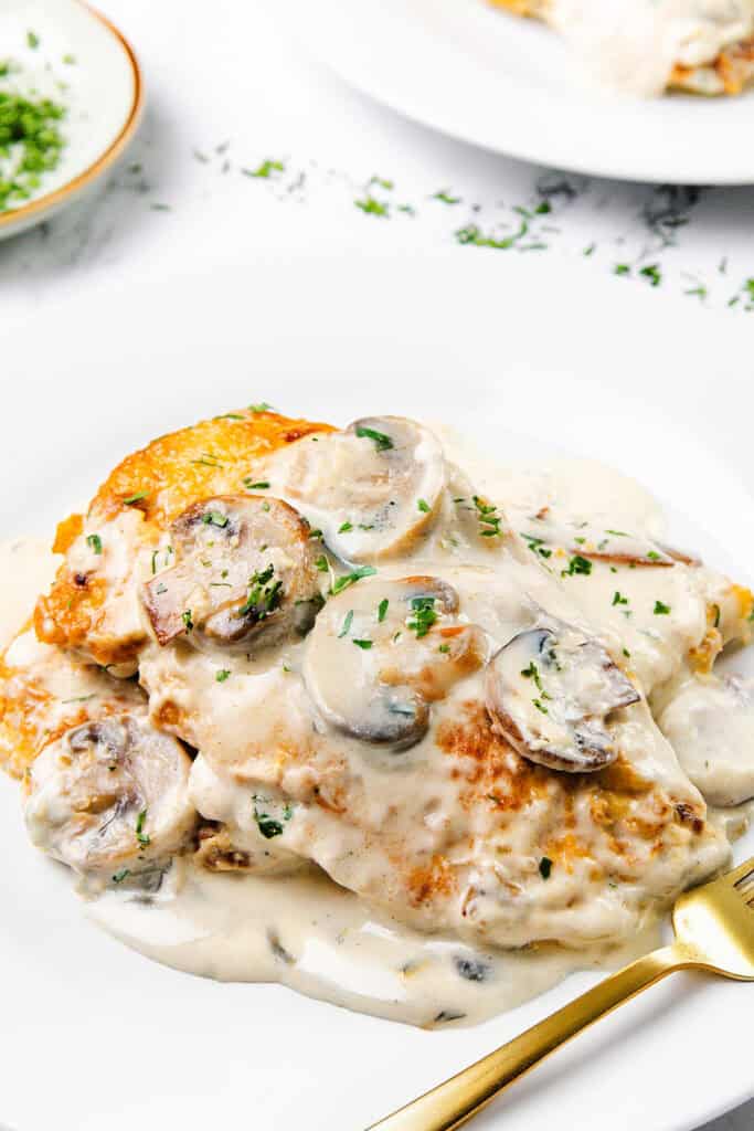 A plate of tender chicken breast topped with creamy mushroom sauce and garnished with chopped herbs, served on a white dish with a gold fork.
