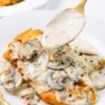 A plate of chicken breasts topped with creamy mushroom sauce, garnished with herbs. A spoon drizzles more mushroom sauce over the chicken. A gold fork rests beside the plate, all on a white background.