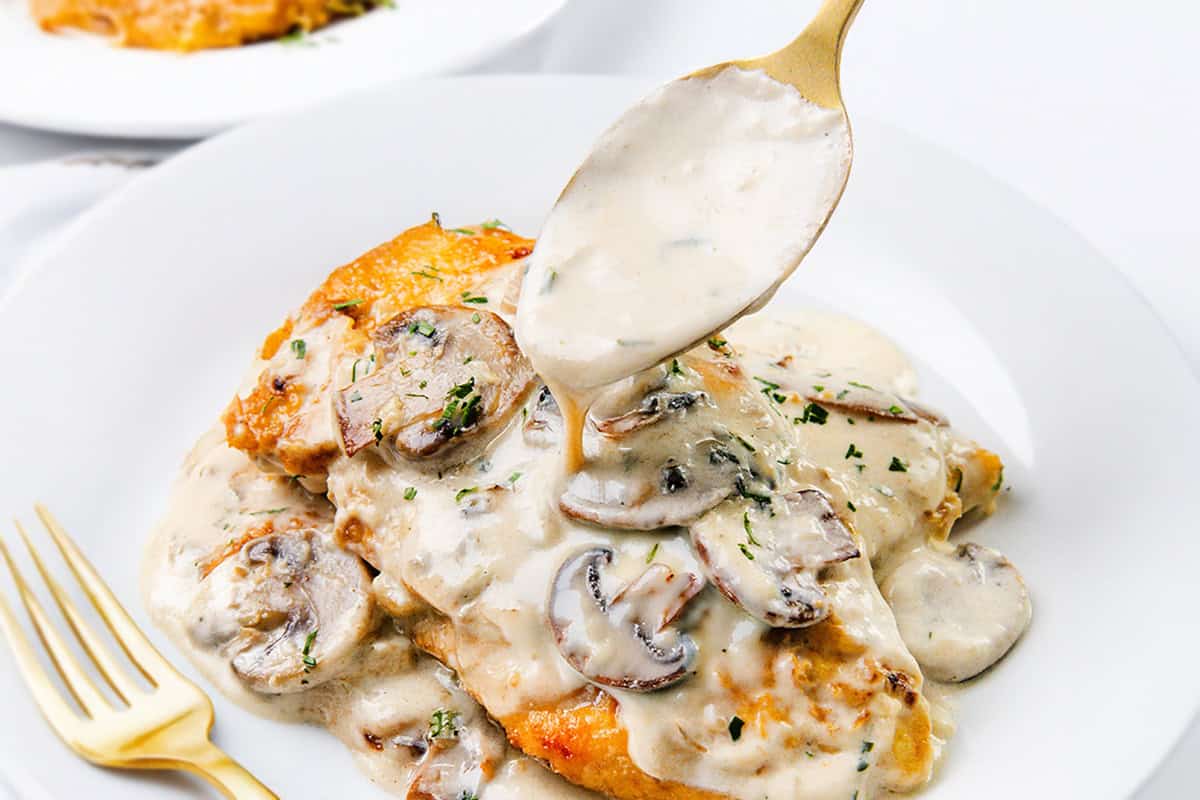 A plate of chicken breasts topped with creamy mushroom sauce, garnished with herbs. A spoon drizzles more mushroom sauce over the chicken. A gold fork rests beside the plate, all on a white background.