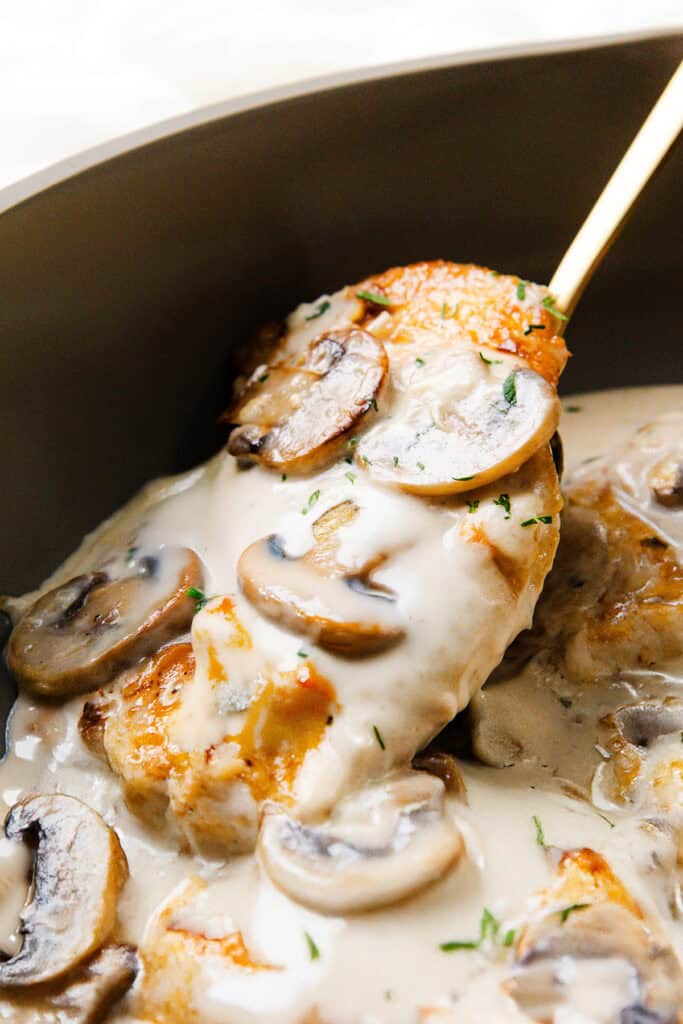 A golden-brown chicken breast is lifted from a pan, covered in creamy mushroom sauce with sliced mushrooms and garnished with chopped herbs.