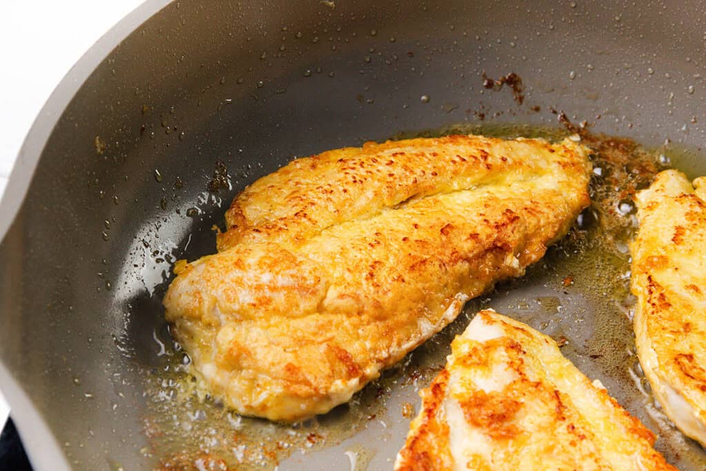 Two pieces of golden-brown fish fillets are being pan-fried in a skillet with oil; the crispy edges and slightly flaky texture make them perfect to pair with a rich, creamy mushroom sauce.