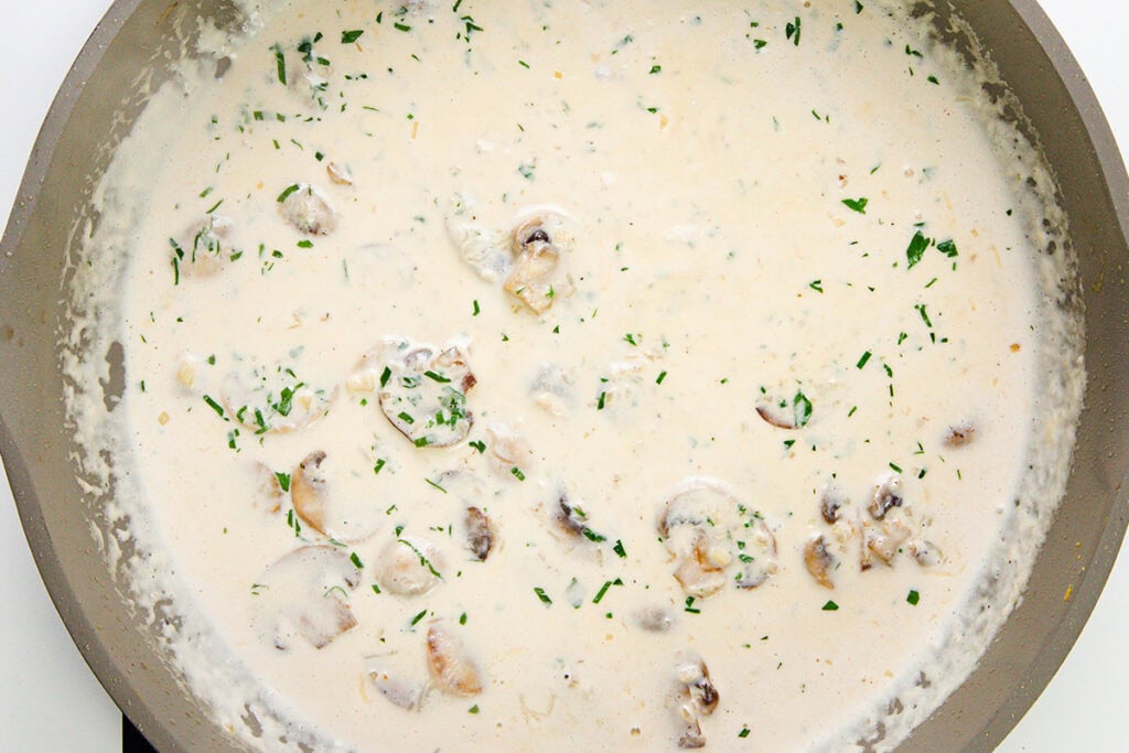 A creamy mushroom sauce with sliced mushrooms and chopped parsley simmering in a pan—perfect for pouring over chicken or pasta.
