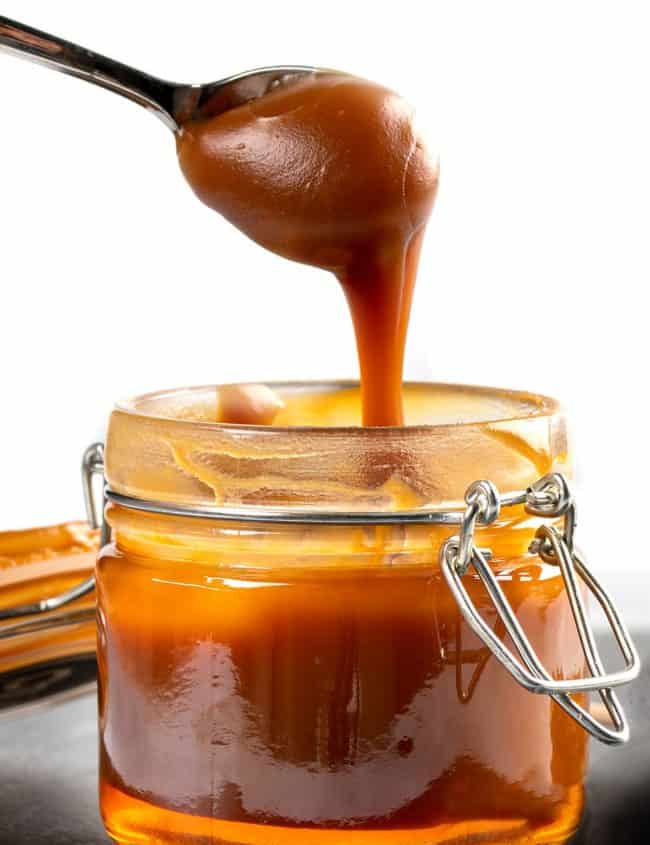 Spoon with tasty caramel sauce over jar on table.