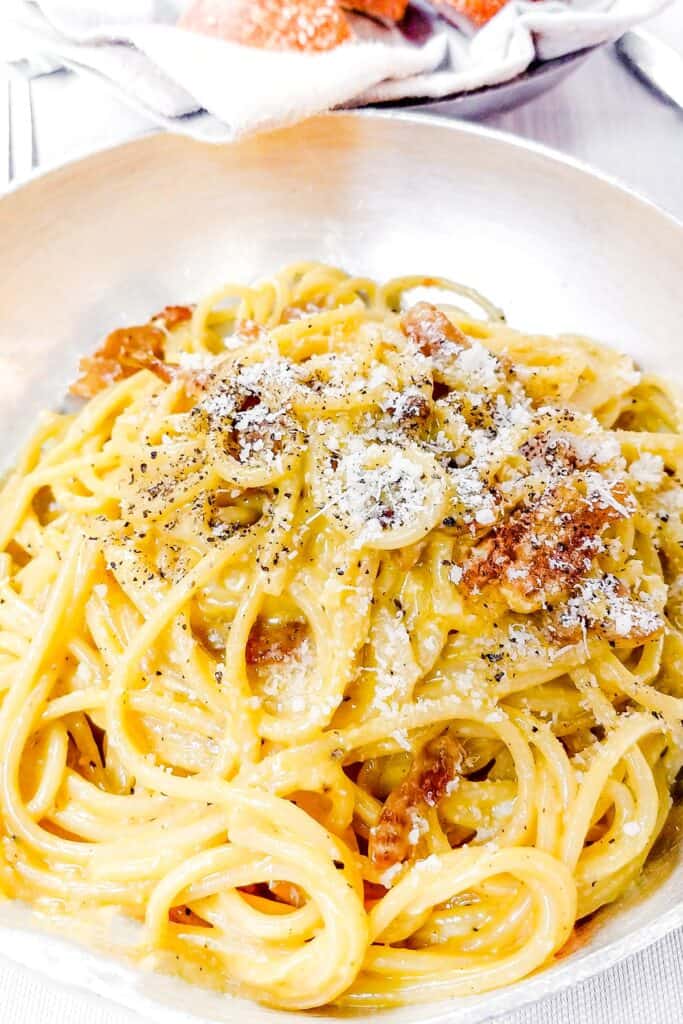 a plate of Spaghetti Alla Carbonara with a basket of bread in the background