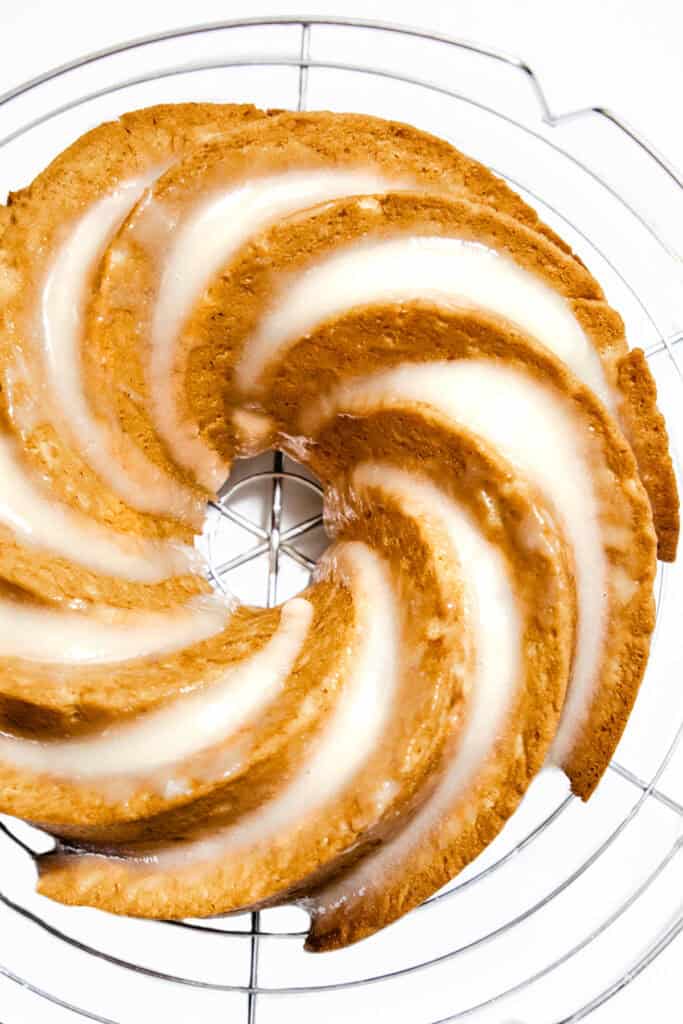 A glazed bundt cake with spiral ridges sits on a round cooling rack. This easy banana cake flaunts a golden brown hue with a shiny white glaze that fills the grooves, creating a striped pattern. The background is white, emphasizing the cake's texture and design.