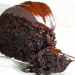 A rich, moist slice of chocolate mayonnaise cake topped with glossy chocolate ganache is placed on a white plate. A small fork rests beside the slice, holding a delectable bite of cake.
