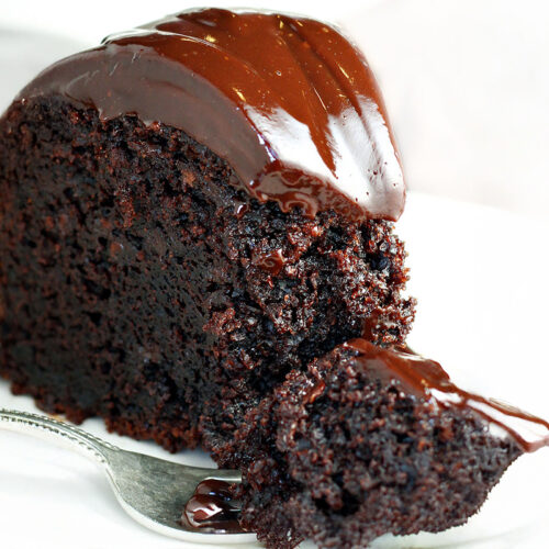 A rich, moist slice of chocolate mayonnaise cake topped with glossy chocolate ganache is placed on a white plate. A small fork rests beside the slice, holding a delectable bite of cake.