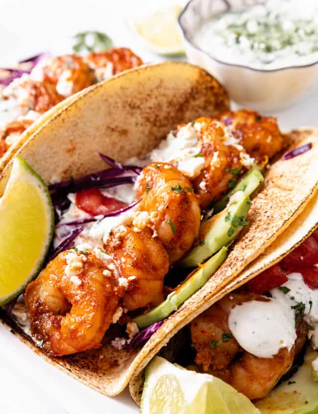 Two shrimp tacos with avocado and red cabbage in corn tortillas, topped with crumbled cheese and creamy sauce, served with lime and extra sauce on the side.