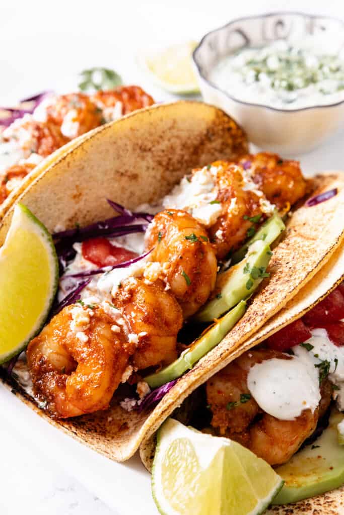 Two shrimp tacos with avocado and red cabbage in corn tortillas, topped with crumbled cheese and creamy sauce, served with lime and extra sauce on the side.