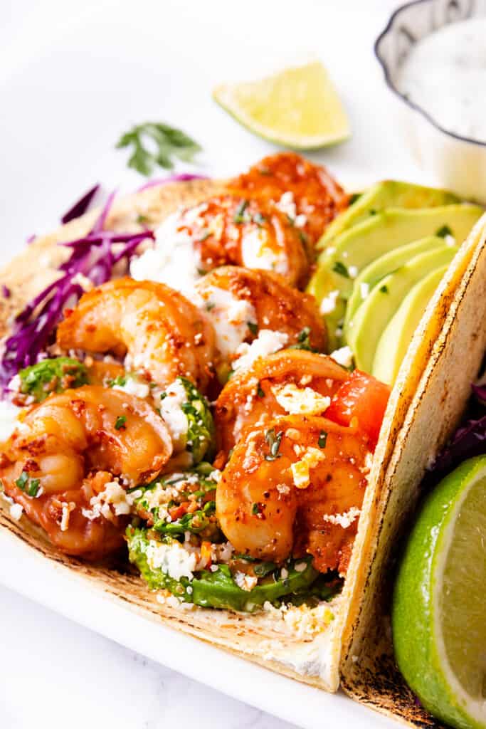 Shrimp tacos with avocado slices and red cabbage in a toasted tortilla, sprinkled with cheese and herbs, served with a side of lime.