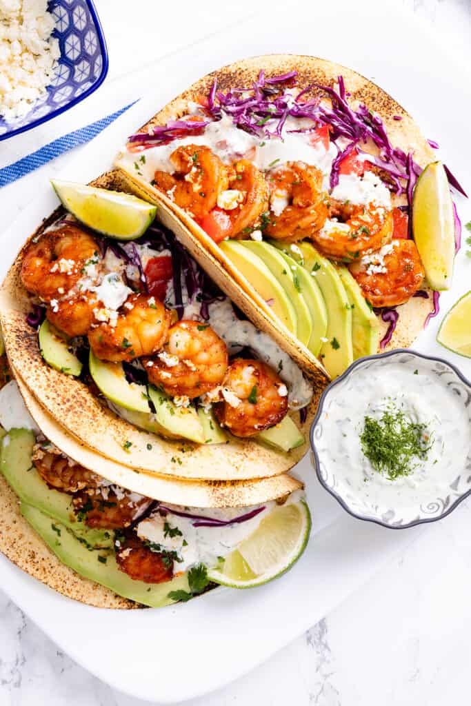 A taco filled with succulent shrimp, nestled among fresh avocado and crunchy red cabbage, topped with crumbled cheese and herbs, accompanied by a lime wedge on the side.