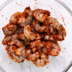 Peeled shrimp coated in a spicy seasoning mix, ready to be cooked, in a clear glass bowl.