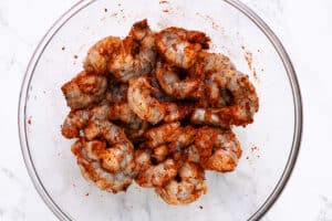 Peeled shrimp coated in a spicy seasoning mix, ready to be cooked, in a clear glass bowl.