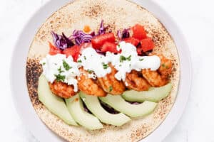 A shrimp taco on a toasted tortilla with avocado slices, red cabbage, diced tomato, and a creamy white sauce, garnished with chopped herbs on a white plate.
