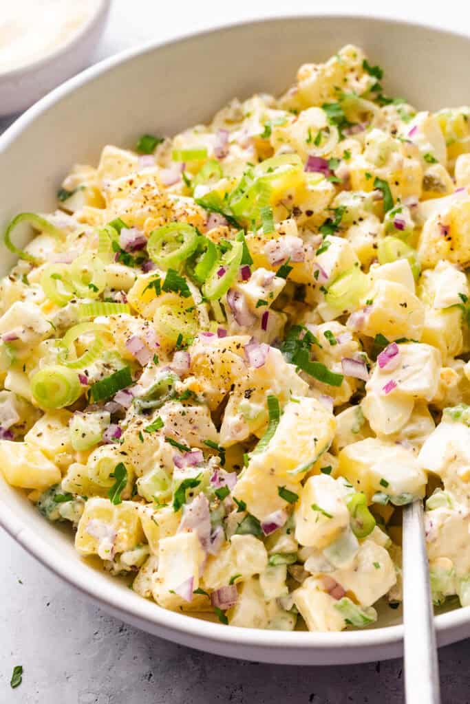 A close-up of homemade potato salad in a bowl, garnished with chopped green onions, red onions, fresh herbs, and black pepper. A serving spoon rests in the creamy salad, ready to serve.
