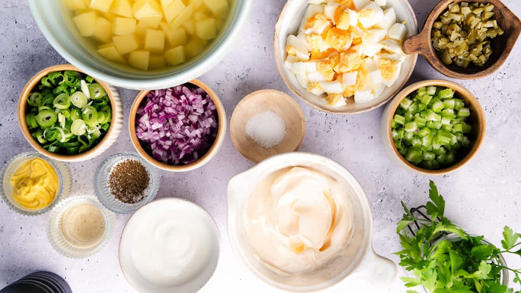 A top-down view of bowls containing chopped potatoes, hard-boiled eggs, pickles, celery, red onion, green onion, parsley, mayonnaise, mustard, spices, and salt on a light surface—everything you need for classic Homemade Potato Salad.